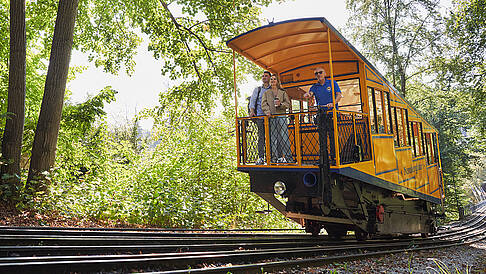 ESWE Verkehr | Nerobergbahn-Fahrt mit Fahrgästen Seitliche Ansicht: Zwei Fahrgäste mit ESWE Mitarbeiter im Nerobergbahn-Waggon während der Fahrt