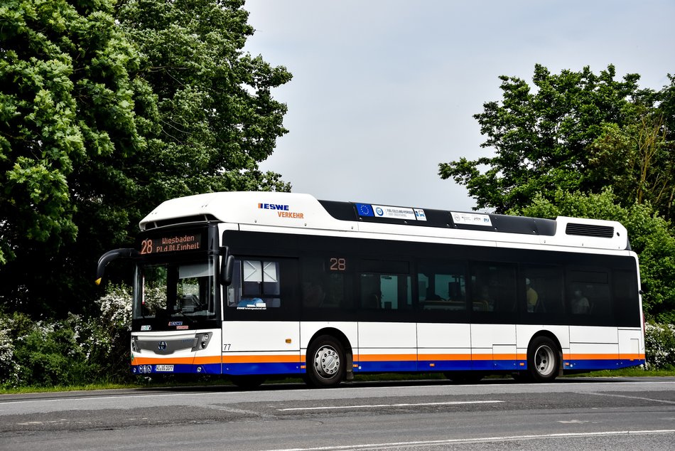 Seitliche Aufnahme eines Brennstoffzellenbus in typischer ESWE-Verkehr-Lackierung auf einer Straße mit zwei Bäumen im Hintergrund.